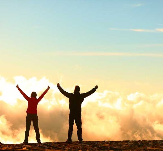 Two hikers raising their arms above the clouds at sunrise